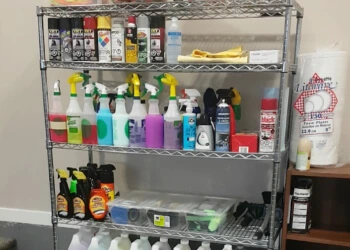 Metal shelving unit stocked with various auto detailing products including spray bottles, aerosol cans, microfiber towels, and gallon jugs, alongside rolls of paper towels.