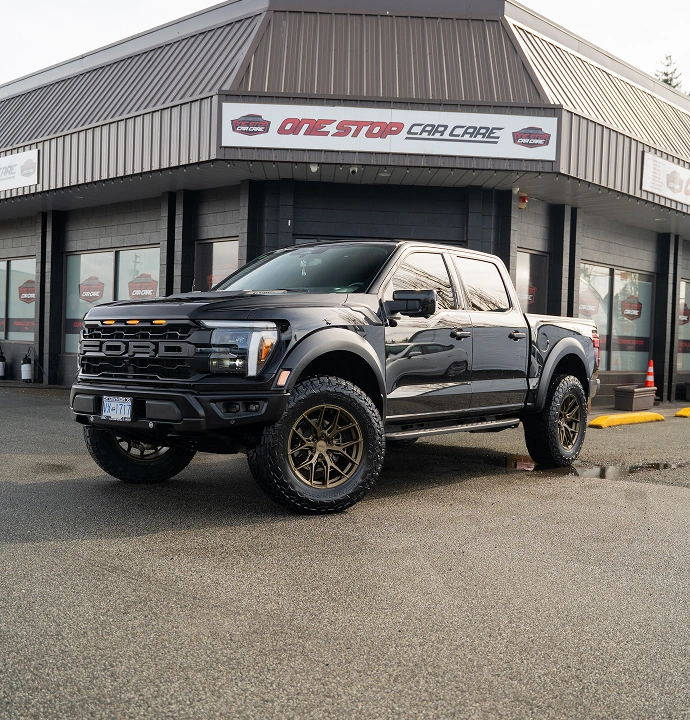 Black Ford Raptor pickup truck with bronze wheels parked in front of the One Stop Car Care building, reflecting a clean and polished finish on a wet pavement.