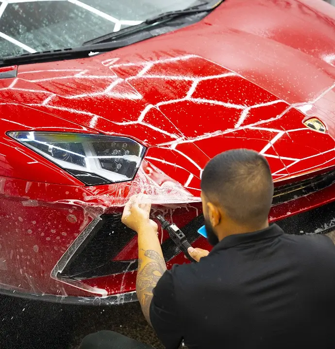 Technician applying a clear paint protection film to the front of a red Lamborghini, smoothing the film over the bumper with a squeegee while the car is covered in soapy water.