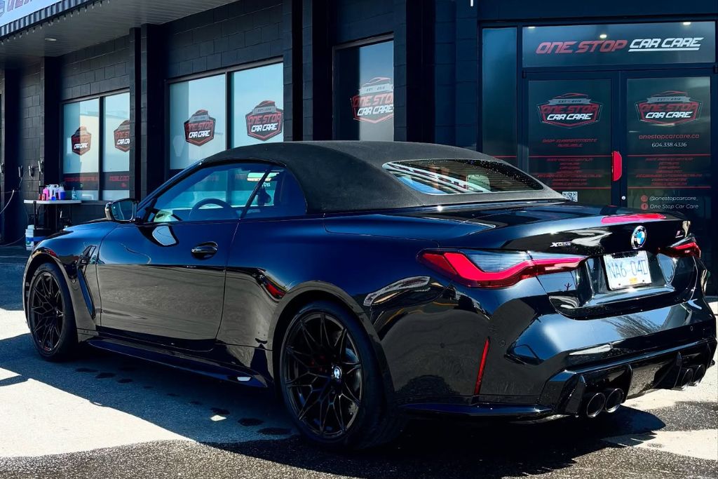 A glossy black BMW convertible with a soft top is parked outside One Stop Car Care, reflecting the bright sunlight and surroundings on its freshly ceramic-coated surface. The detailing shop’s logo is prominently displayed on the building's windows and doors in the background.
