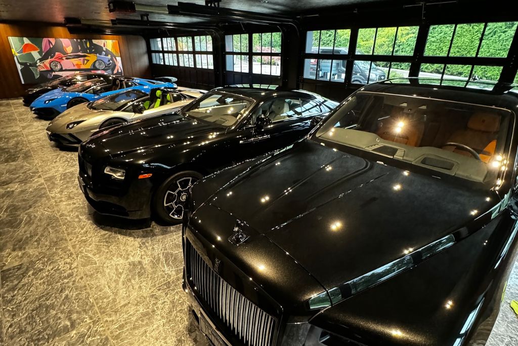 A row of luxury vehicles including Rolls-Royces and Lamborghinis parked in a polished showroom with bright overhead lighting reflecting off their pristine ceramic-coated surfaces.