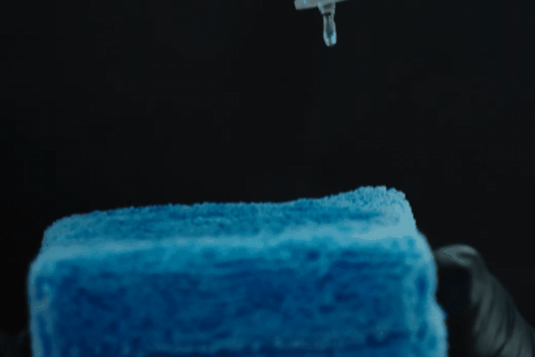 Close-up of a gloved hand holding a blue microfiber applicator while a drop of ceramic coating solution drips onto it from a dropper against a black background.