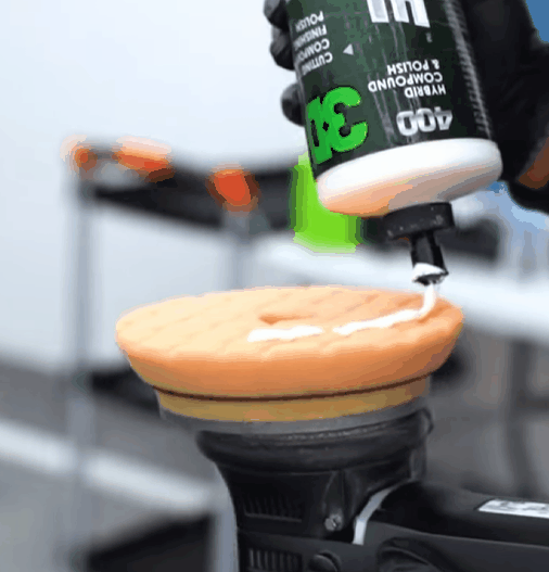 A gloved hand squeezes hybrid compound polish labeled “3D 400” onto an orange foam polishing pad attached to a power buffer in a clean detailing shop.