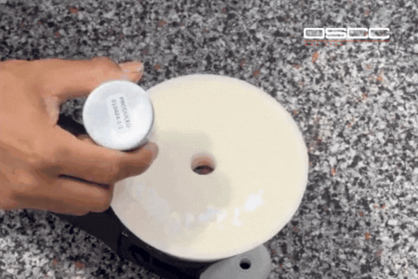 A hand squeezes ceramic compound onto a white polishing pad attached to a buffer tool, preparing for paint correction or surface treatment. The OSCC (One Stop Car Care) logo appears in the corner of the frame.