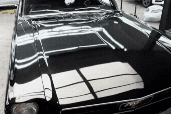 Animated pan over a glossy black classic Ford Mustang front end showing the hood and chrome grille under bright garage lights. The rotating view highlights sharp reflections and a smooth surface. The continuous motion emphasizes the deep shine achieved by a professional ceramic coating.