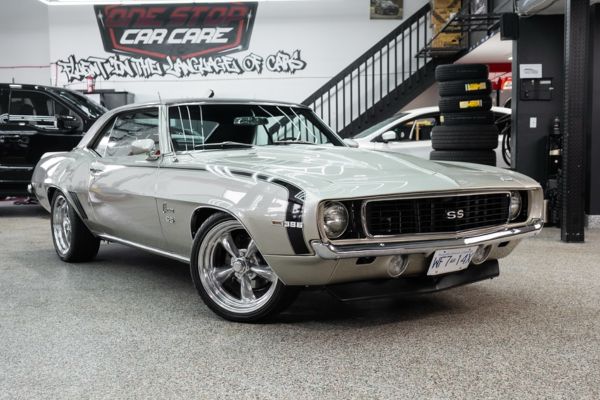 Classic silver Chevrolet Camaro SS parked inside a detailing garage under bright lights. Graffitied One Stop Car Care sign hangs above a black metal staircase with stacked tires nearby. Reflections on the hood and wheels highlight the flawless ceramic coating.