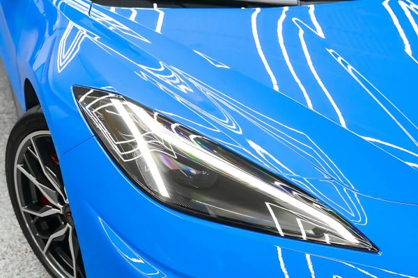 Blue sports car front end shows a sleek headlight and hood reflecting bright workshop lights. High gloss finish highlights the protective ceramic coating.