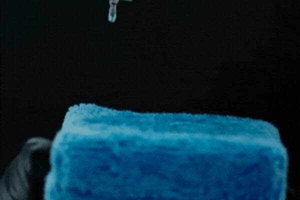 Close-up of a gloved hand holding a blue microfiber applicator as clear ceramic coating drops onto it from a glass dropper, illustrating the precise application process. This visual supports the topic: what is ceramic coating, with a focus on its professional use in Surrey.