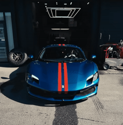 A glossy blue Ferrari with red racing stripes sits halfway out of a detailing garage, showcasing a flawless ceramic-coated finish. Nearby equipment and a controlled workspace highlight One Stop Car Care’s professional installation environment.