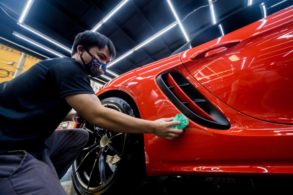 A detailing professional wearing a face mask applies ceramic coating with a green applicator pad to the side panel of a shiny red sports car under bright workshop lights. This image visually supports the discussion around what is ceramic coating and its professional application in Surrey.