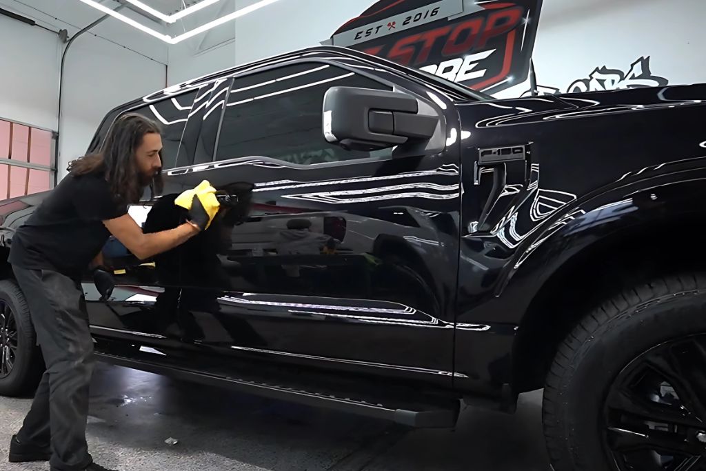 A technician wearing gloves hand-applies ceramic coating with a yellow microfiber cloth to the side of a glossy black pickup truck inside a detailing shop. This step illustrates the hands-on application phase in the ceramic coating process, answering what is ceramic coating for Surrey vehicle owners.