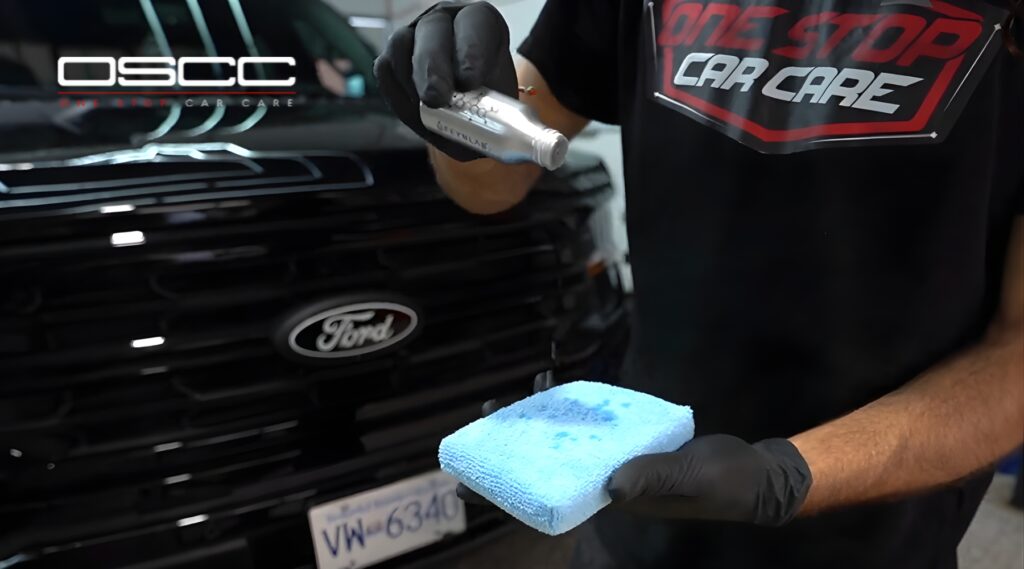 A car detailing technician in Surrey, BC holds a blue microfiber applicator pad while pouring ceramic coating solution from a silver bottle, with a black Ford vehicle in the background. This visual supports the topic: what is ceramic coating.