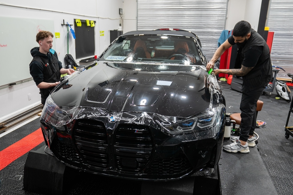 Two One Stop Car Care technicians apply paint protection film to the front of a black BMW inside the Surrey detailing studio, carefully smoothing the material over the hood and fenders to ensure long-term durability and warranty-backed protection for high-end vehicles.