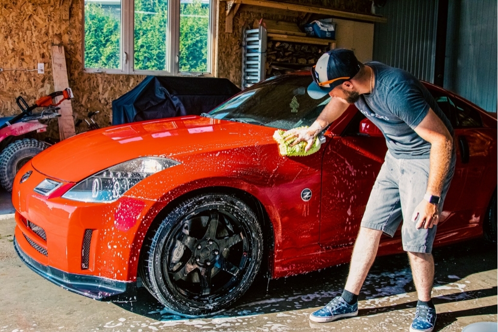 A man hand-washes a bright red sports car with a microfiber mitt in a well-lit garage, following proper paint protection film care practices. This visual supports PPF maintenance tips for keeping the film clean and effective.