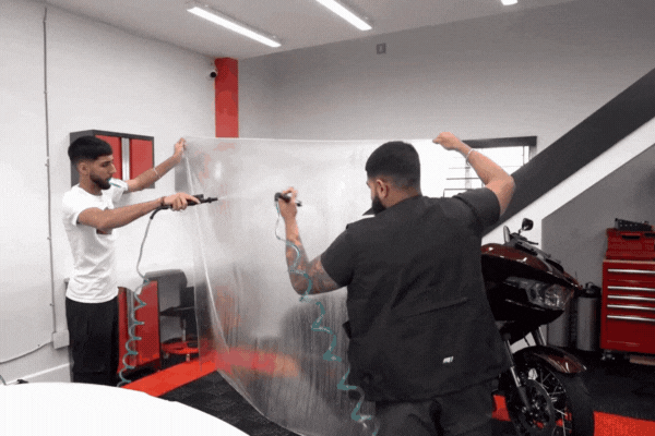 Two technicians at One Stop Car Care in Surrey, BC, stretch a clear sheet of paint protection film across a surface, showcasing the Power of PPF as a strong, flexible barrier for vehicle paint protection in a professional detailing shop.