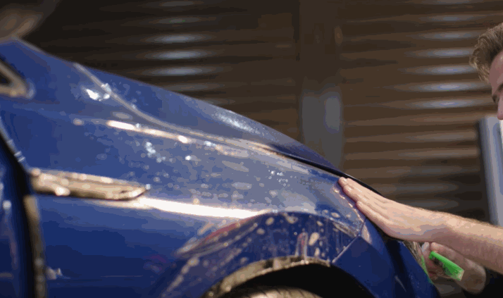 A man carefully applies blue paint protection film on the side of a car, pressing it down with a squeegee to remove water and air.