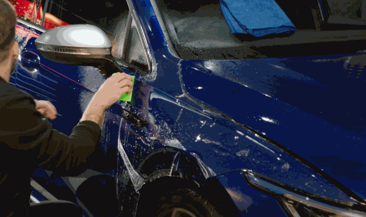 A technician uses a green squeegee to apply paint protection film on the front side of a blue SUV, smoothing out the film with water for adhesion.