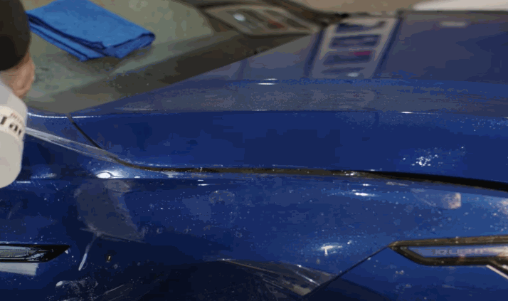 A close-up of a person peeling off the liner from clear paint protection film on the hood of a blue car, revealing a smooth, shiny surface underneath.