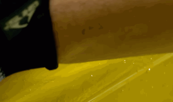 A close-up of a hand using a squeegee to smooth out yellow paint protection film on a car surface. The squeegee glides along the film, pushing out air bubbles.