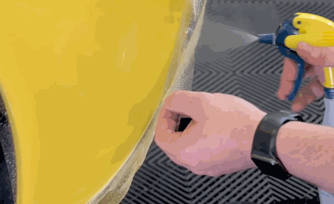 Close-up of a person applying paint protection film to a yellow car hood using a spray bottle to position the film smoothly.