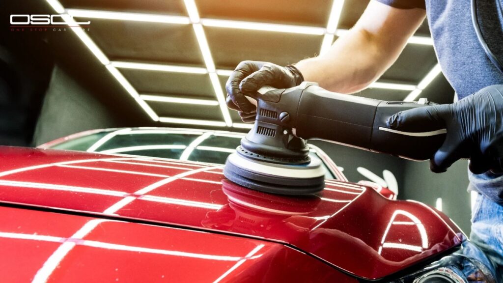 Auto detailer using a rotary polisher on the hood of a red car under bright grid lighting during the ceramic coating process.