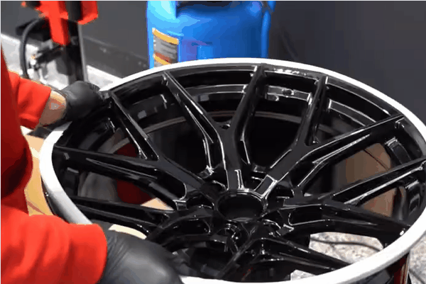Close up of a technician in a red uniform and black gloves carefully handling a glossy black alloy wheel in an auto shop. The image supports a guide about choosing the right wheels and tires in Surrey by showing professional inspection and care.