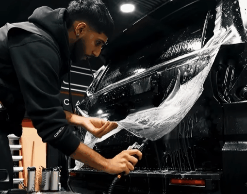 Alt text: A technician applies paint protection film to the front bumper of a black vehicle, pulling the clear film into place while working on the wet surface by hand. This supports content about why paint protection film is worth it for Surrey drivers who want to protect paint, maintain appearance, and preserve resale value.