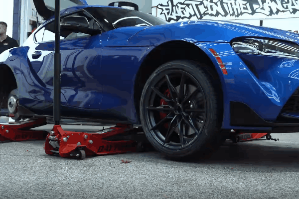 Blue sports car raised on red hydraulic jacks inside an auto shop, with the front wheel and low profile tire prominently shown to support the section about why comfort changes when wheel size changes. The image emphasizes how larger wheels are often paired with shorter sidewalls that can create a firmer ride, and a graffiti style wall mural appears in the background with text that is not fully legible.