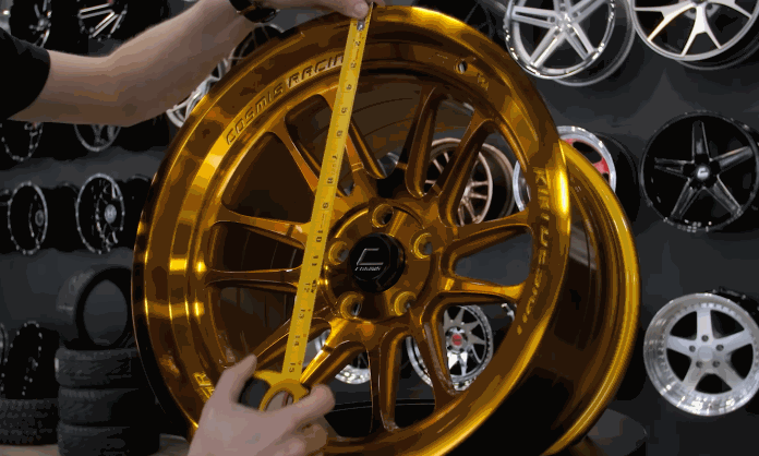 A technician measures the rim size of a glossy gold wheel with a yellow tape measure. More display wheels hang on the wall behind it.