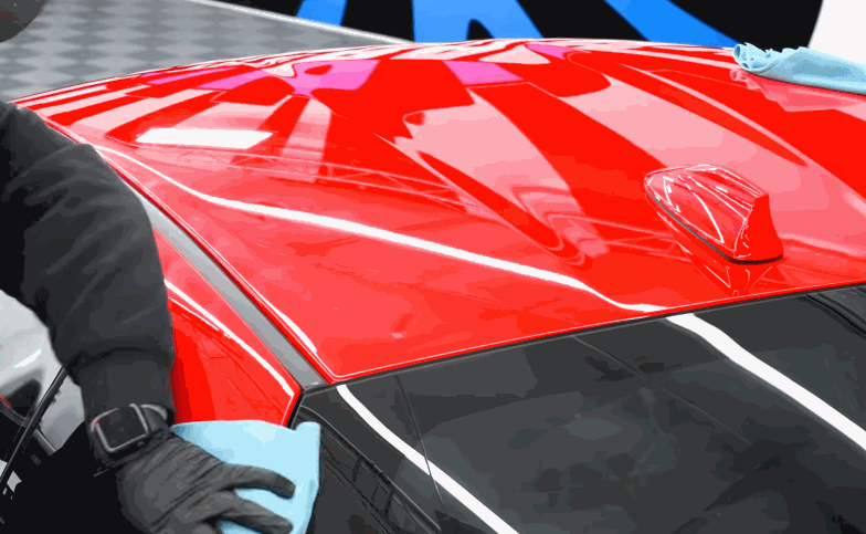 A technician wipes the red roof of a vehicle with a blue microfiber towel during ceramic coating prep. The glossy surface reflects overhead lights across the painted panel.