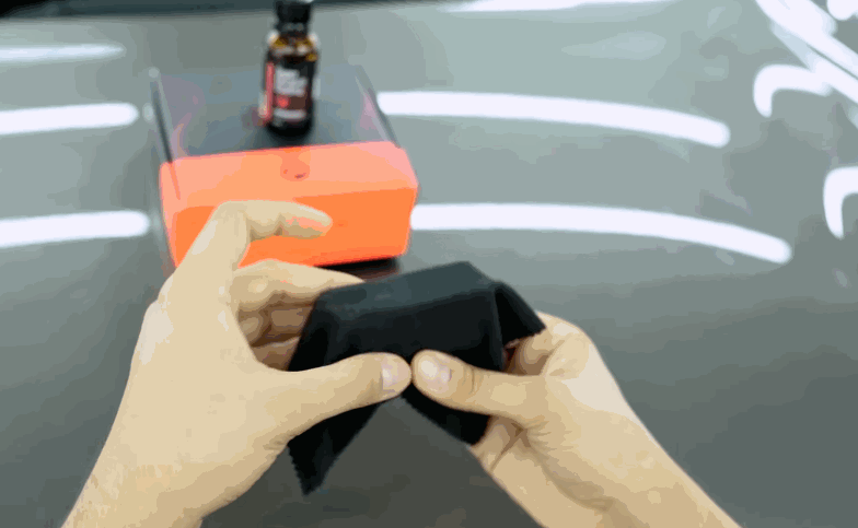 Hands prepare a ceramic coating applicator cloth on a glossy gray vehicle hood. A small coating bottle and orange applicator block sit nearby for the next step.