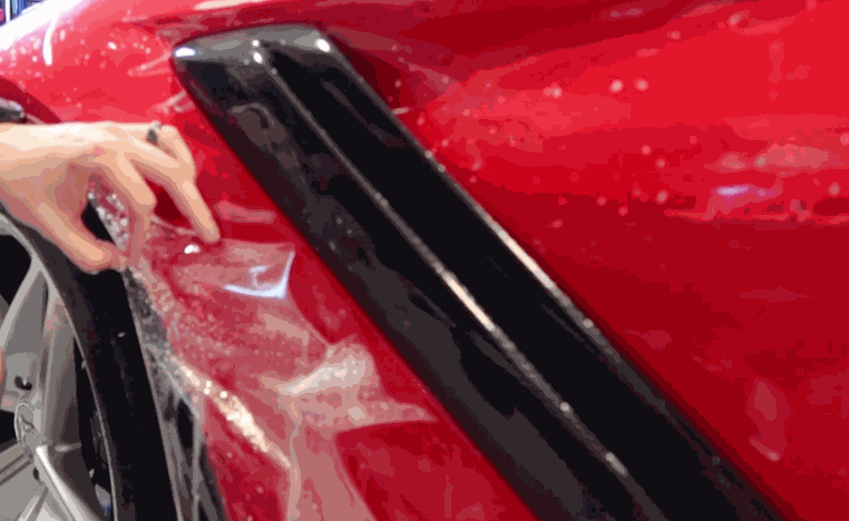 A technician smooths paint protection film onto the side panel of a red vehicle near the wheel arch and black trim. The clear film is being aligned over the wet painted surface.