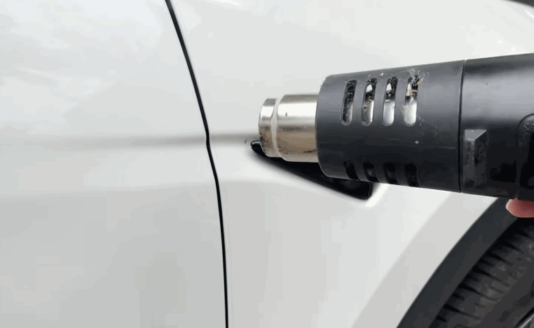 Heat gun warming the edge of paint protection film near a trim piece on a white car.