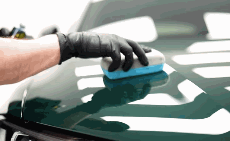 Gloved hand moves a blue applicator pad across a glossy green car hood before ceramic coating.
