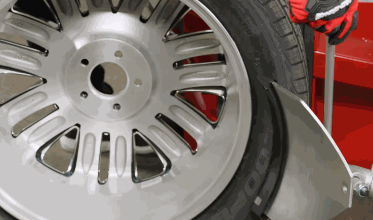 Tire installation machine presses a tire onto a silver wheel with a red vehicle in the background.
