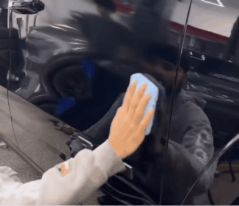 Detailer gently wiping a black vehicle's glossy door panel with a blue microfiber cloth during professional ceramic coating aftercare.