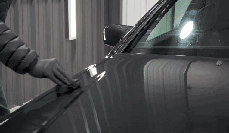 Detailer wearing a black glove wipes the gray body panel of a car under bright detailing lights before ceramic coating application. The smooth surface reflects the overhead lighting clearly.