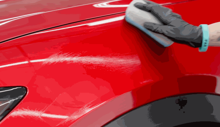 Detailer uses an applicator pad across the red front fender of a car while applying ceramic coating. The glossy paint reflects the surrounding lights as the product is spread over the surface.